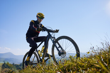 Fototapeta na wymiar Man in safety helmet and glasses cycling uphill on sunny day with blue sky on background. Male bicyclist in cycling suit climbing uphill on mountain bike. Concept of sport and active leisure.