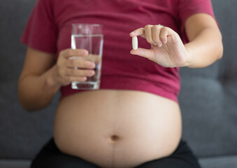happy smiley young pregnant Asian woman sitting on a couch holding a glass of pure water and pill. Pregnancy healthy and people concept