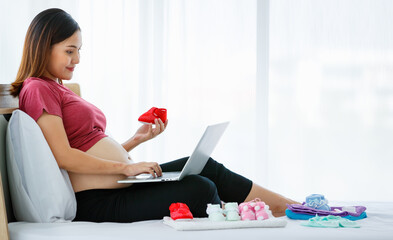 portrait of young active Asian pregnant woman sitting on a bed holding a cute baby shoe while enjoy using and looking at a laptop doing shopping online