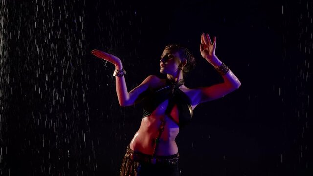 oriental dance of young woman, dancer lady is performing movements under rain in darkness