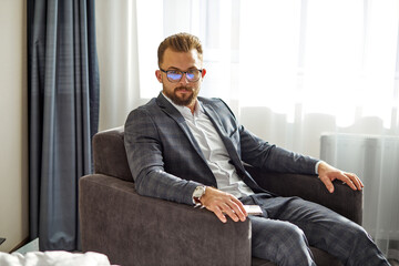 male in spectacles and in suit look at camera sitting on couch in hotel room, before meeting, conference