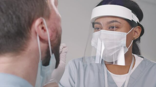 Tilting-up closeup with slowmo of female african american nurse in protective mask and face shield taking throat swab from adult man for covid-19 test