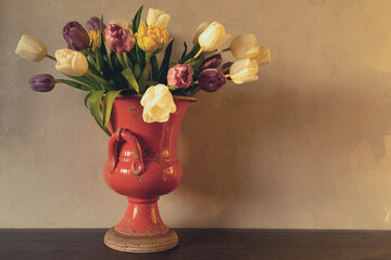 Fresh spring beautiful bouquet of multicolored tulips in a red beautiful ceramic vase in a room on a shelf against the wall