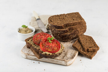 Black bread sandwich with tomatoes and hummus on a wooden board on a light background.