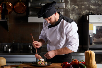 Chef adding piquancy to dish, finishing with flavouring of meal on plate, wearing cap and apron, cook alone in restaurant kitchen - obrazy, fototapety, plakaty