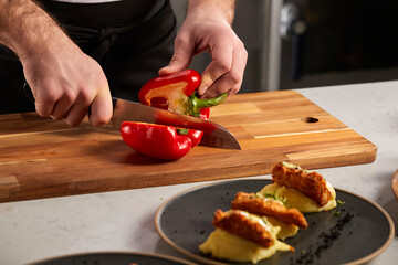 Slicing sweet red pepper on wooden cutting board, professional cook preparing salad. Female hand cuts capsicum with knife. restaurant staff at work. close-up