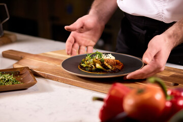 Unusual tasty dish on black plate represented by professional cropped cook in apron, plate on wooden board, fresh yummy meal