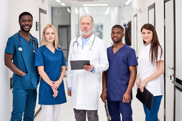 Confident doctor in white uniform and young doctors therapists posing looking at camera, elderly man holding clipboard in hospital, multidisciplinary medical team in white coats in aisle