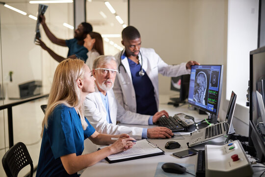 Diverse Doctors and Radiologists Discuss Diagnosis while Looking at Computer Tomography X-ray image Brain Scans Results, Discussing. Focus On Blond Nurse sitting At Desk