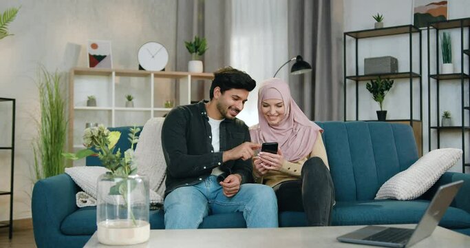 Pleasant happy smiling muslim couple in love sitting one near one on comfortable sofa at home and watching interesting videos or apps on mobile