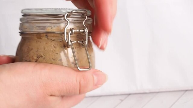 A glass jar with chicken liver pate is opened and held by women's hands.