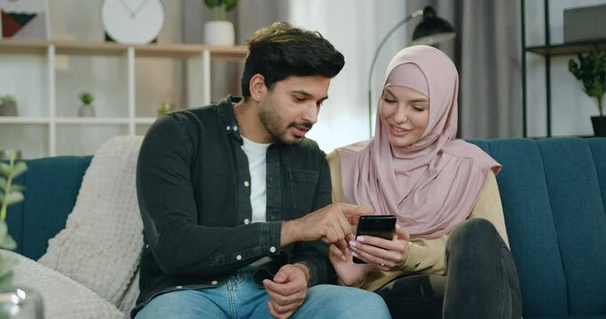 Good-looking loving happy smiling muslim couple relaxing on comfortable sofa and revisioning favourite photos on mobile while spending leisure at home