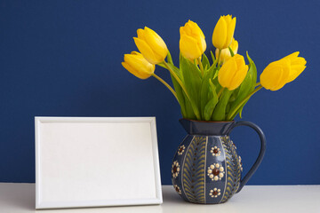 ceramic jug with yellow tulips and white frame on blue background