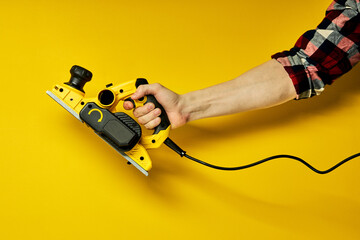 cropped male hands carpenter holding working woodworking power tool in hands, on yellow background. Close up of instrument, copy space
