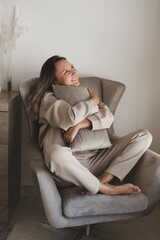 Happy and smiling woman dressed in comfy loungewear sitting in grey armchair and holding a decorative pillow. Coziness and comfort at home. Casual outfit - 419330747
