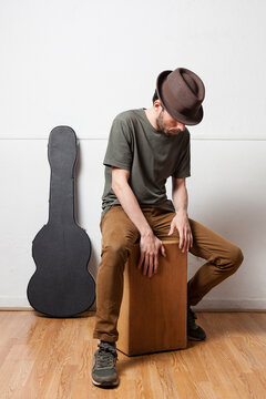 White spanish man wearing hat playing peruvian cajon on a white background with guitar case and wooden floor. Flamenco passion concept.