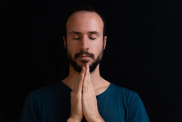 young relaxed man meditating on black background with closed eyes. pensive man with beard holding hands together, concentrated doing yoga