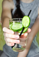 A girl holds detox water in a glass with sliced cucumbers. Blurry.