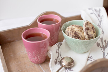 Homemade round gingerbread cookies with icing in a ceramic bowl and two mugs of tea with tea strainer on a wooden tray with a napkin