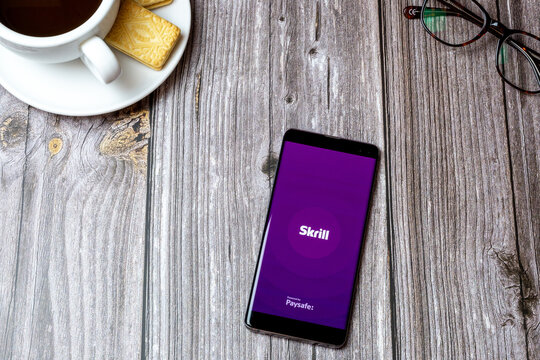 02-24-2021 Portsmouth, Hampshire, UK A mobile phone or cell phone on a wooden table with the Skrill app open next to a coffee and glasses
