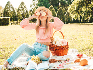 Obraz premium Young beautiful hipster woman in trendy summer jeans, pink T-shirt and hat. Woman making picnic outside. Positive model sitting on plaid on grass, eating fruits and cheese, holding lemons in hands
