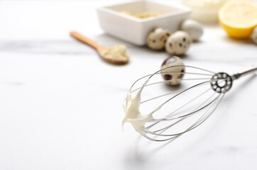 Metal whisk and white tasty homemade dressing for salad, eggs, lemon, other ingredients on the white table.Empty space