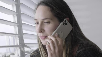 woman talking on the phone while looking out the window. Lady speaking with friends on a smart cellphone. looking out to the street from the shutters light on beautiful face. chatting with a colleague