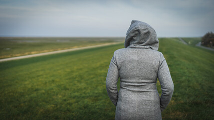 Frau in grauer Jacke geht einsam und alleine auf dem Deich spazieren