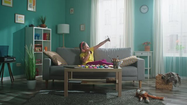 Small funny girl is sitting on sofa in living room and taking selfie, trying to entertain during quarantine, Zoom in, Slow motion.