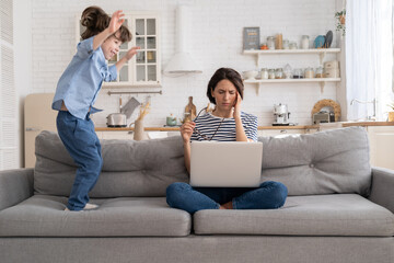 Irritated mother sitting on couch working on laptop at home, distracted from noise and touching her temple, hyperactive little kid jumping on sofa and drawing attention. Tired mom can't concentrate.