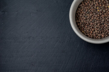 top view of a fragment of a white ceramic bowl filled with fresh lentils on the top right edge of a dark slate surface that is the background of the frame and serves as a copy space.