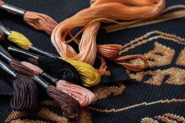 Multicolored skeins with floss threads scattered on black canvas covered with cross stitch pattern...