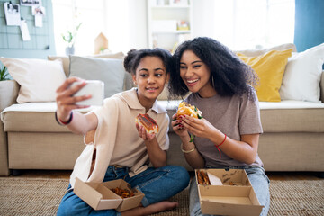 Portrait of young sisters with burgers indoors at home, taking selfie.