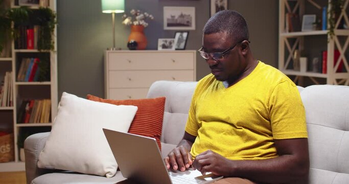 Good-looking man in glasses resting on couch and using laptop in evening. Adult Afro American checking e-mail, receiving good news and rejoicing at home. Education, business, job concept.