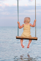 Blond girl sitting on rope swing over the water. Back view. Happy childhood.