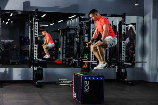 A focused athlete in an orange T-shirt and gray shorts jumps on the gym box in a darkened fitness studio with a mirror. Sports lifestyle, healthy life - Powered by Adobe