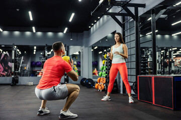 A woman and a man in sportswear throw a fitness ball in the gym. The man is in a squat position and...