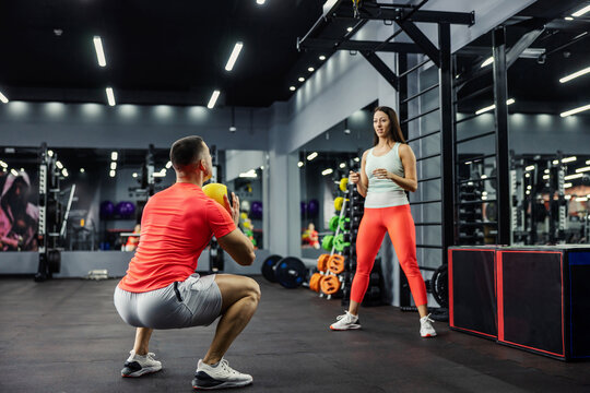 A Woman And A Man In Sportswear Throw A Fitness Ball In The Gym. The Man Is In A Squat Position And Is Getting Ready To Throw The Ball To The Girl Who Is Standing Upright.Sports Challenge, Couple Goal