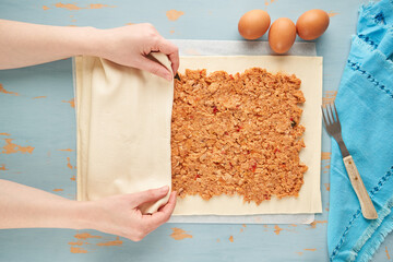 A person places dough over the tuna, tomato, onion and bell pepper filling to prepare an empanada on a blue wooden table. Tuna pie. Typical Galician dish from Galicia and Spain