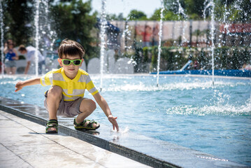 Little boy plays in the square near pool with water jets in the fountain at sunny summer day. Active summer leisure for kids in the city