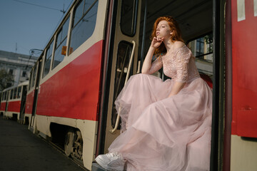 Amazing beautiful red-haired young woman in retro style fancy prom pink dress sitting on steps of old old red Soviet tram. Creative concept. Public transport fashion. rough sneakers shoes.  pensively  © Iryna&Maya