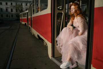 red-haired young beautiful girl sits on the steps of an old red Soviet tram. kitsch style fashion prom style fancy pink retro dress high rough sneakers shoes. Eastern Europe public transport. 