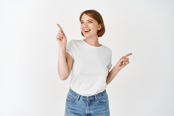 Happy beautiful female pointing sideways but looking left with cheerful smile, making choice, picking things on shopping, standing against white background