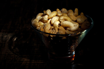 Cashews in a clear glass bowl on an old shabby board. Nuts on a brown wooden table. Contrasting dramatic light as an artistic effect.