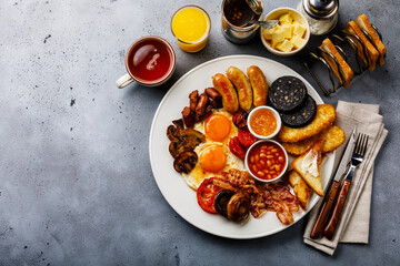 Full fry up English breakfast with fried eggs, sausages, bacon, black pudding, beans, toasts and tea on gray concrete background