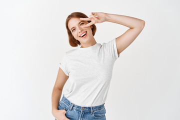 Cheerful young woman with short hair, showing v-sign over eye, standing positive and happy, white background