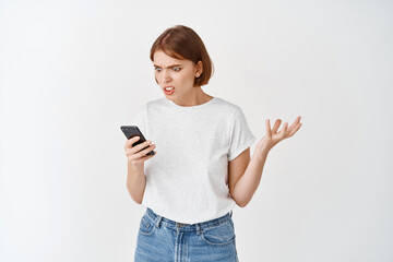 Annoyed girl look at smartphone screen and complaining, staring angry at display with raised hand,...