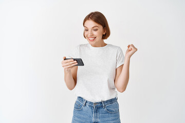 Excited woman winning video game on smartphone, looking happy at smartphone screen and say yes,...