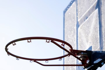 A portrait of an outdoor basketball rim without a net and a frozen plexiglass backboard with blue...