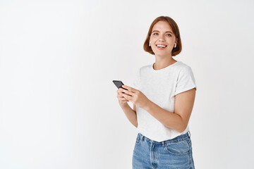 Portrait of beautiful happy girl holding smartphone screen and smiling, listening music in wireless headphones, standing against white background